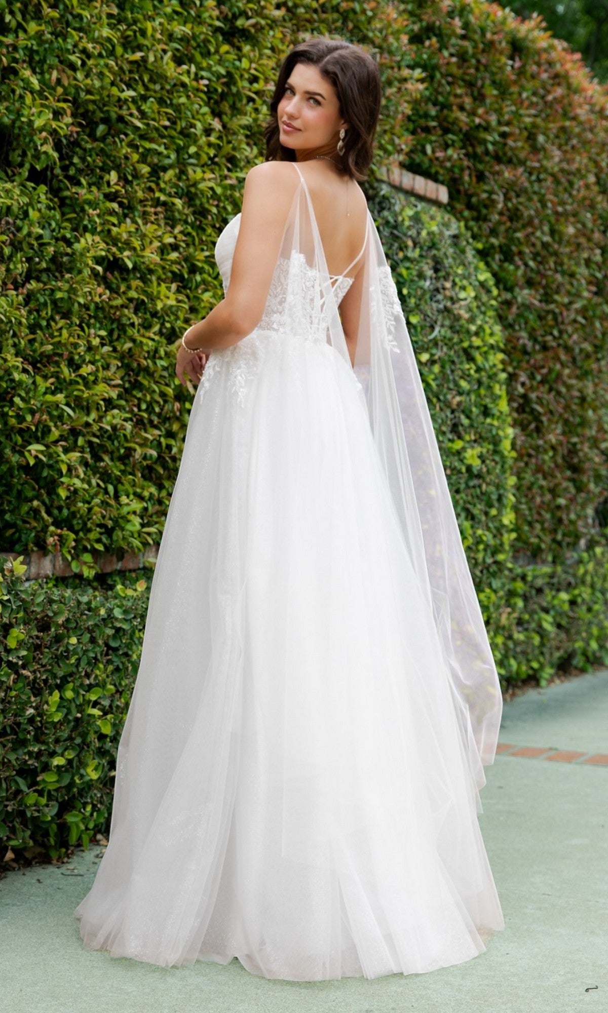 A woman stands outdoors in front of a green hedge, looking over her shoulder while wearing the Cinderella Couture 4217 long formal dress—a white, sleeveless bridal gown featuring pearl-beaded lace details and a flowing tulle A-line skirt.